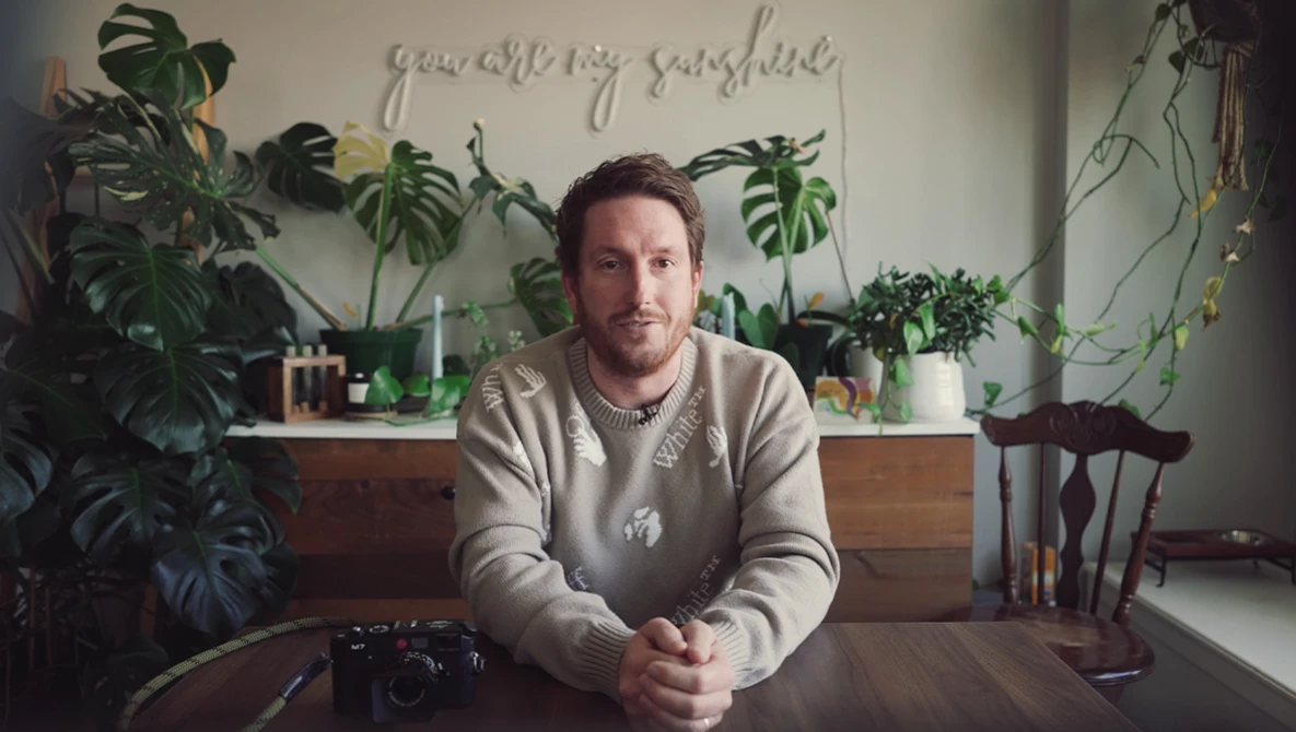 Man seated at wooden table with camera, surrounded by potted plants and natural window light.