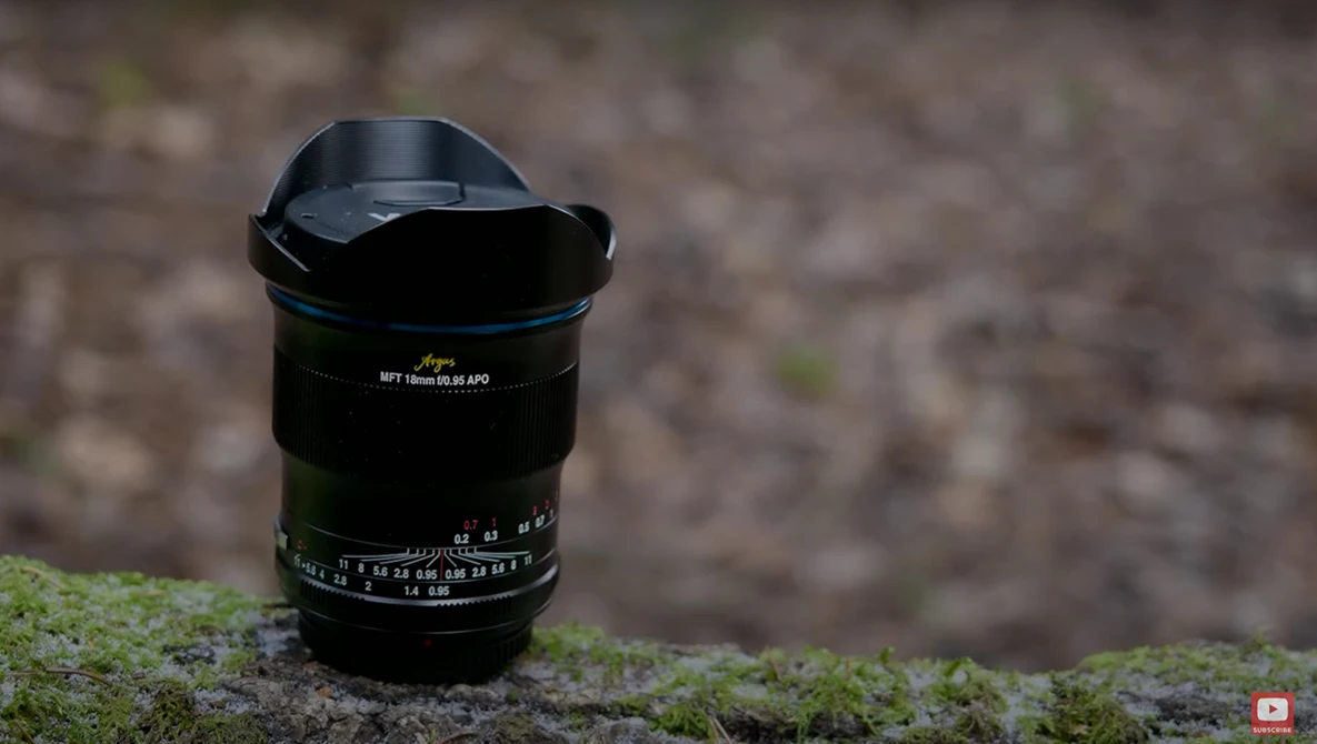 Wide-angle lens with lens hood mounted on moss-covered ground against blurred brick wall.