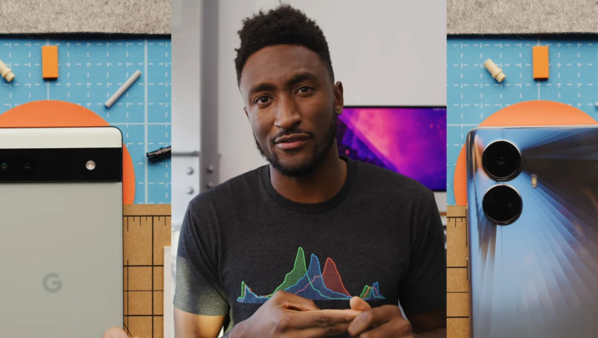 Man in graphic t-shirt sitting in front of smartphone and tablet displays in a tech-focused studio setting.