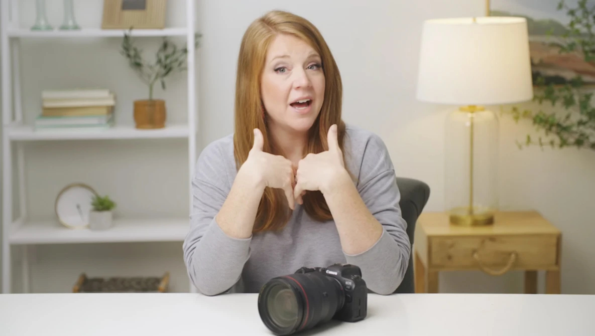 Woman with reddish-brown hair gesturing enthusiastically toward a professional DSLR camera on a white desk.