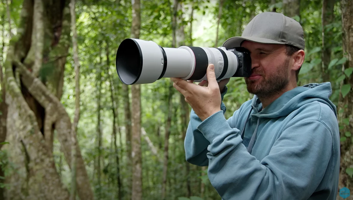 Photographer with telephoto lens in forest setting, wearing gray cap and blue jacket.