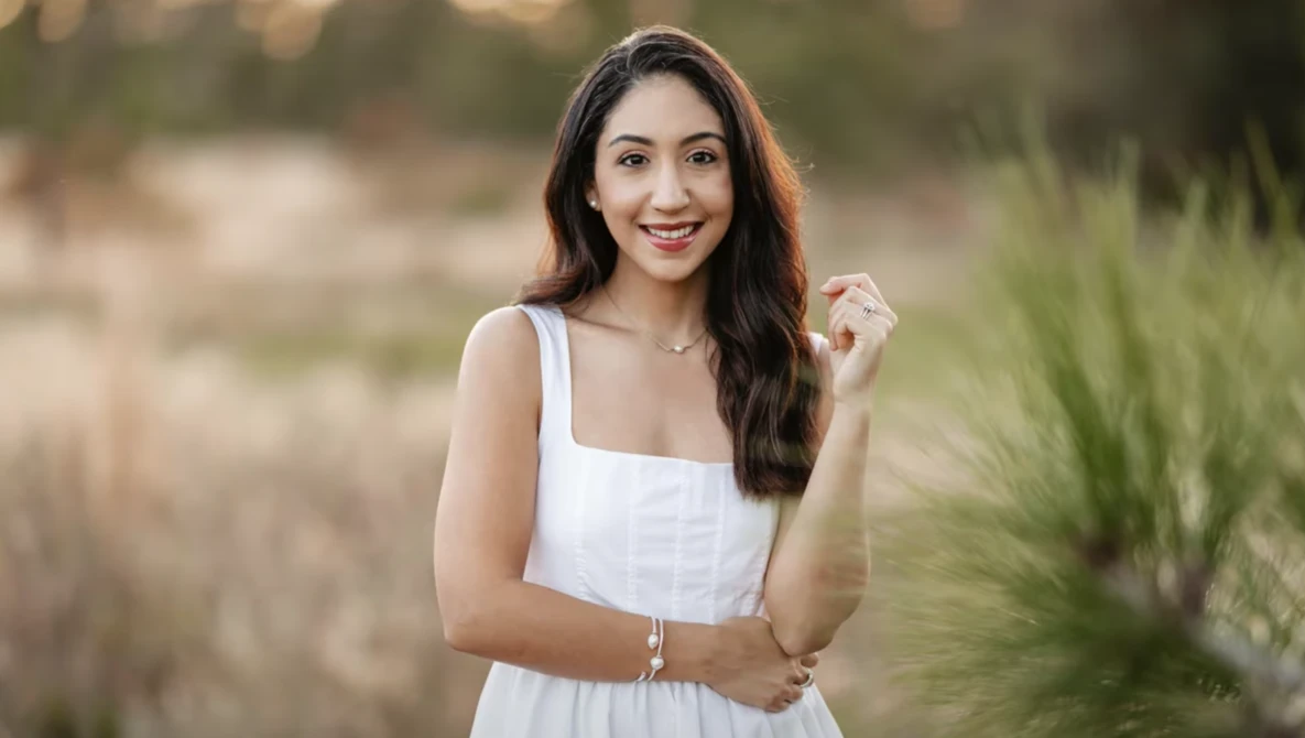 Woman in white dress smiling at camera in outdoor setting with blurred natural background.