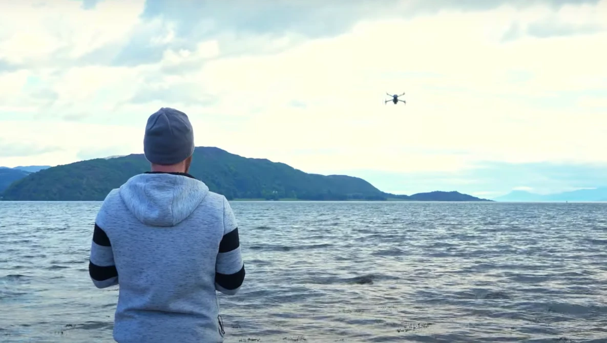 Person in blue and black hoodie standing in shallow water, operating a drone hovering over the ocean.