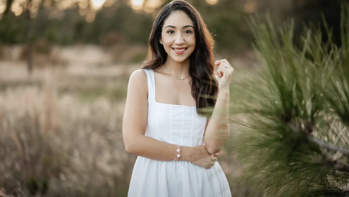 Woman in white sleeveless dress posing outdoors during golden hour with natural vegetation in background.