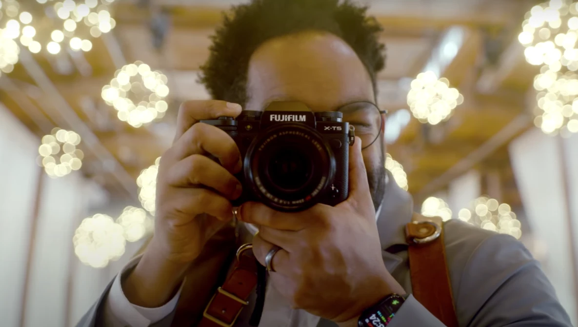 Photographer holding a Fujifilm camera up to their face, with warm bokeh lights blurred in the background.