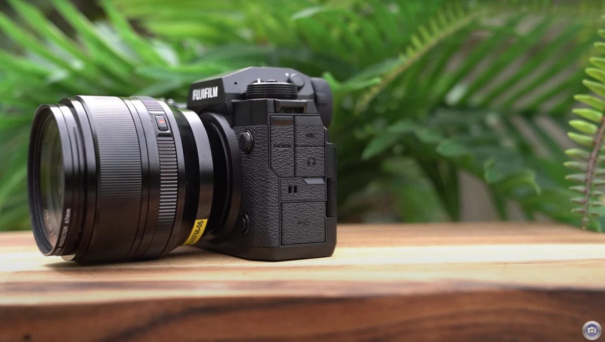 Fujifilm mirrorless camera with attached lens on wooden surface, green foliage in background.
