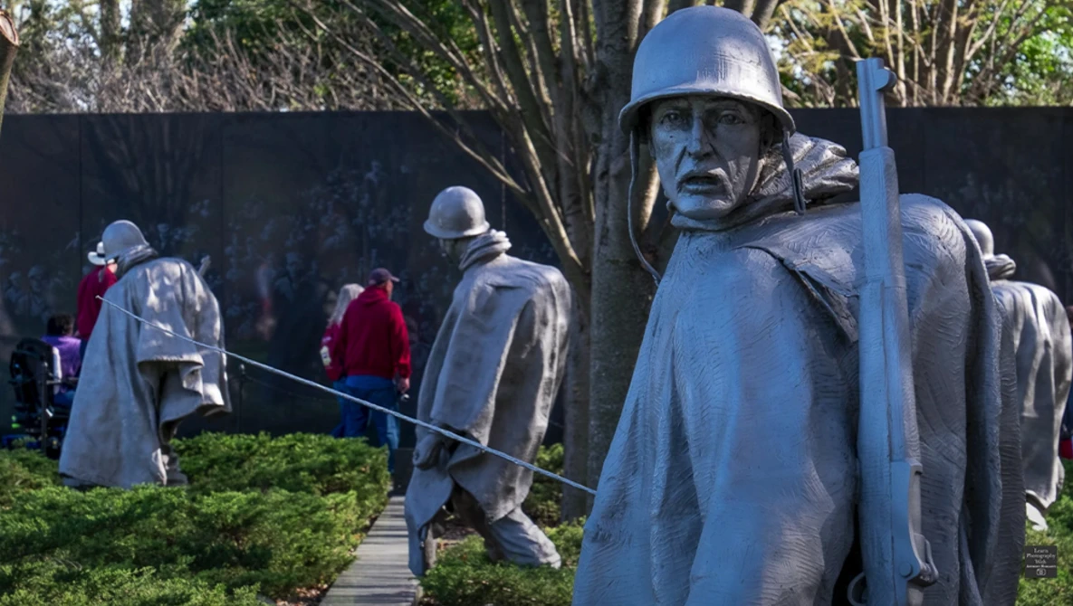 Stone statues of soldiers in military uniforms and helmets standing in a memorial garden with visitors walking in the background.