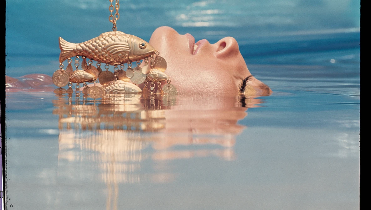 Person submerged in shallow water with ornate golden fish sculpture suspended above their face.