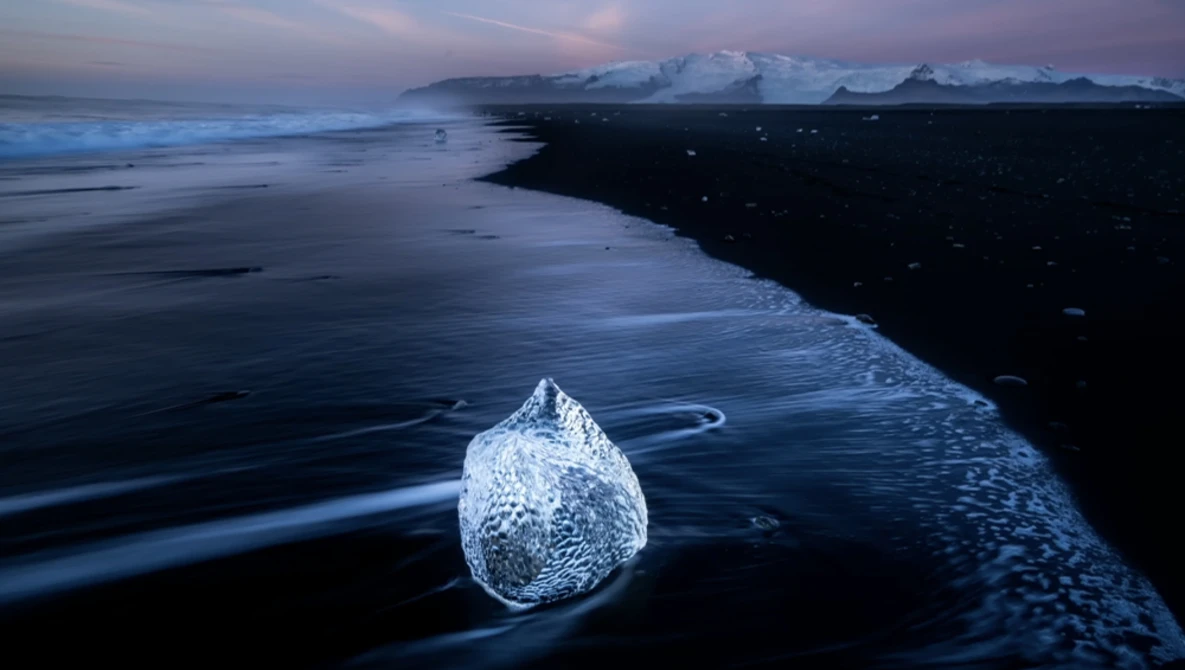 Translucent ice chunk on dark volcanic sand beach during twilight hour.