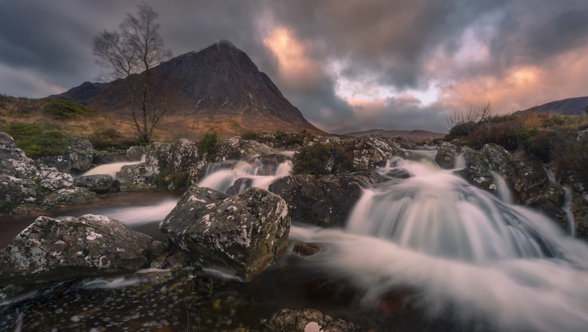 Rushing mountain stream flowing around moss-covered rocks beneath a dramatic peak at sunset.