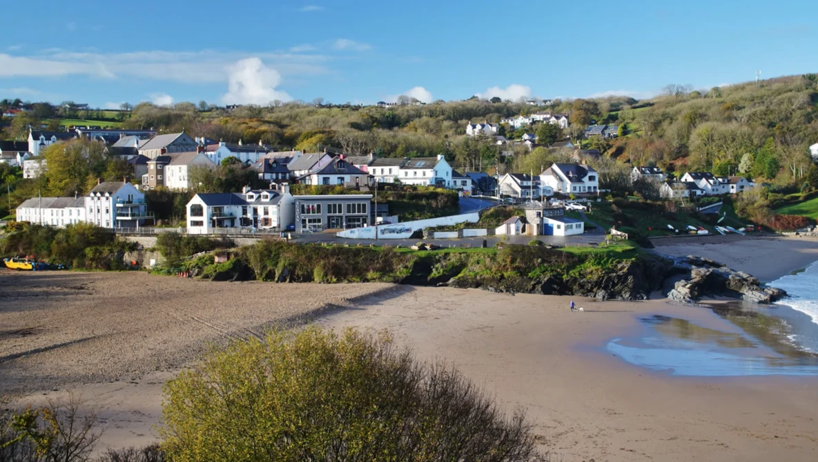 Coastal village with sandy beach and residential buildings backed by forested hillside under clear sky.