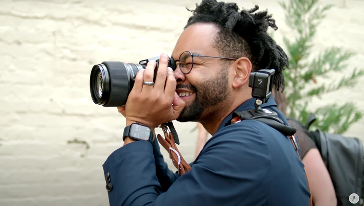 Photographer with glasses and dreadlocks holding a DSLR camera to his eye while smiling.