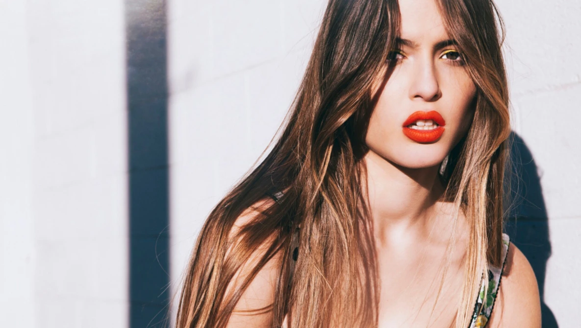 Woman with long brunette hair and red lipstick against a white wall with dramatic shadow.