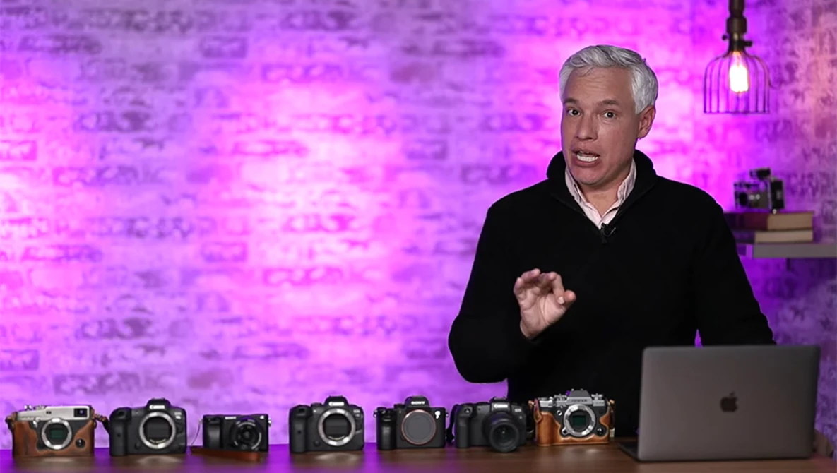 Man in black jacket gestures while discussing seven cameras arranged on table in front of him.