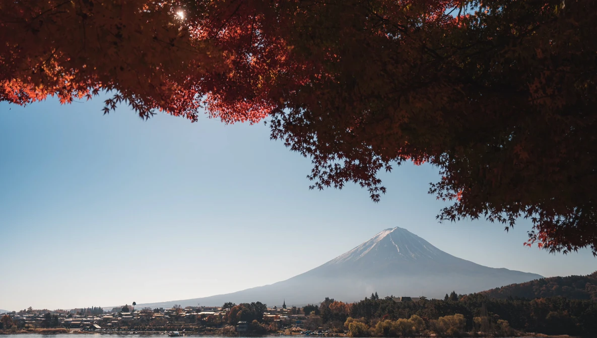Snow-capped volcanic mountain framed by red autumn foliage in foreground, with clear blue sky and lakeside town below.
