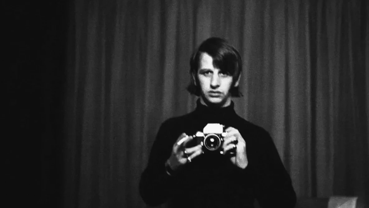 Man in dark sweater holding vintage camera directly at lens against curtained backdrop.
