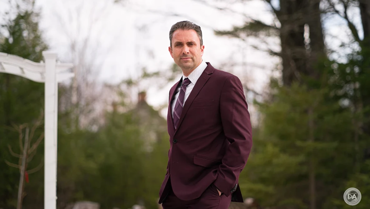 Man in burgundy suit standing outdoors with evergreen trees in background.