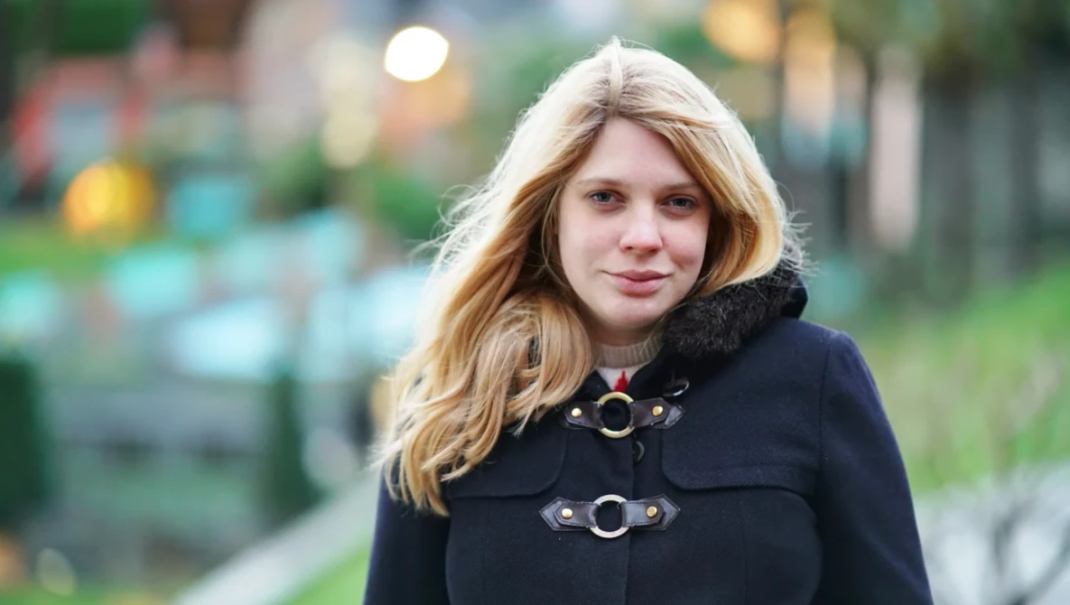 Woman with long blonde hair wearing a black coat with decorative buckles, photographed outdoors with blurred background.
