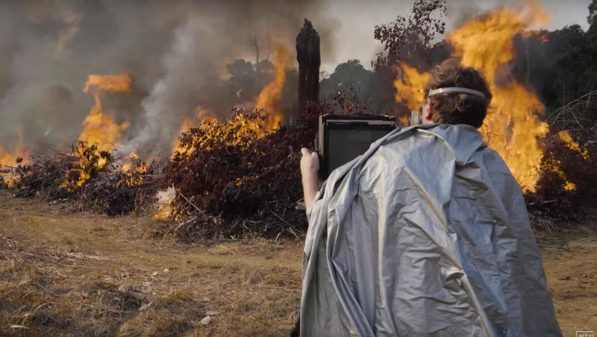 Person in light-colored clothing operating equipment while managing large outdoor fire with intense flames.