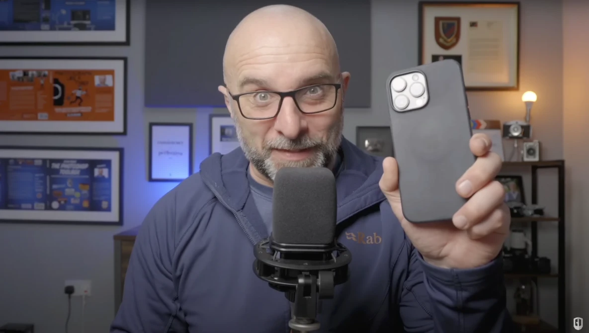 Male presenter holding a smartphone with camera lenses visible, speaking into a studio microphone.