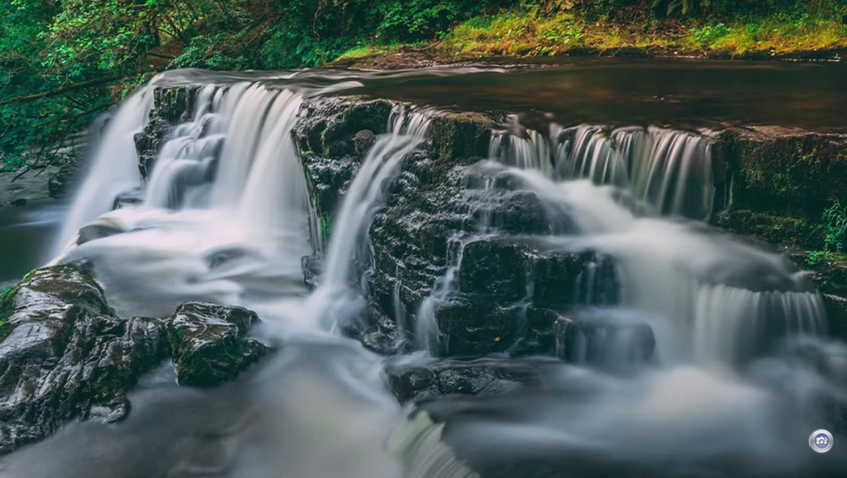 Cascading waterfall flowing over layered rock formations surrounded by dense green forest.