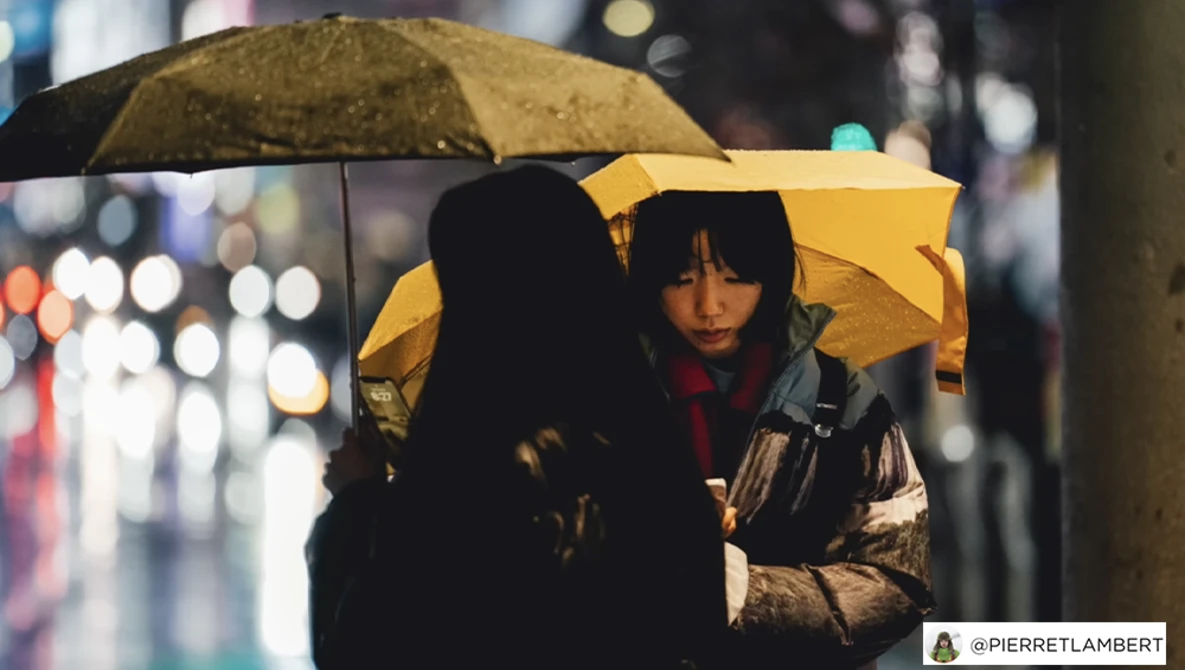 Two people under yellow umbrellas at night on a wet street with blurred city lights in the background.