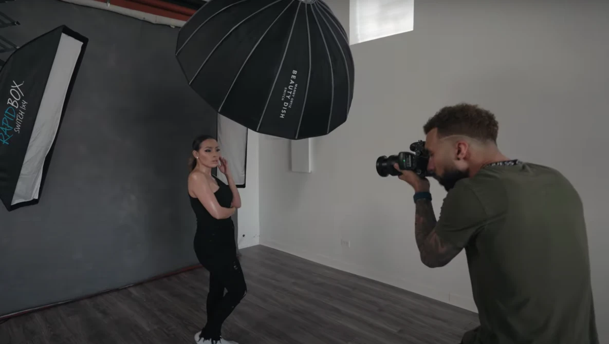 Photographer with camera on tripod shooting portrait of woman in black clothing against gray backdrop with studio lighting setup.