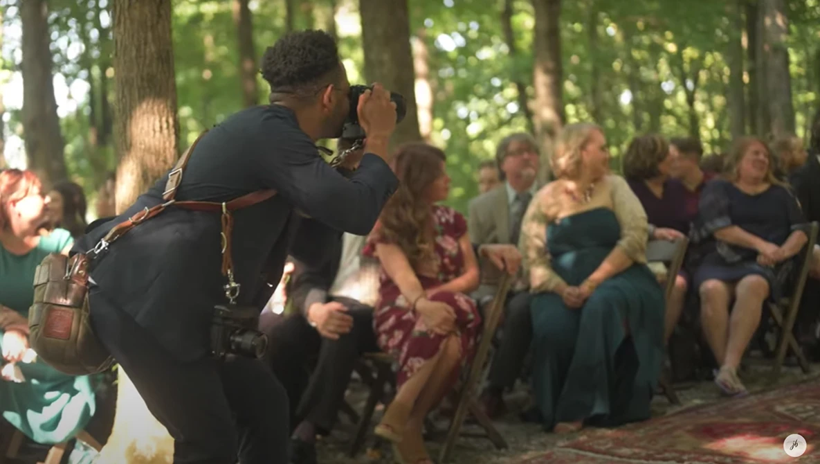 Photographer with camera on shoulder capturing an outdoor forest ceremony with seated guests in background.