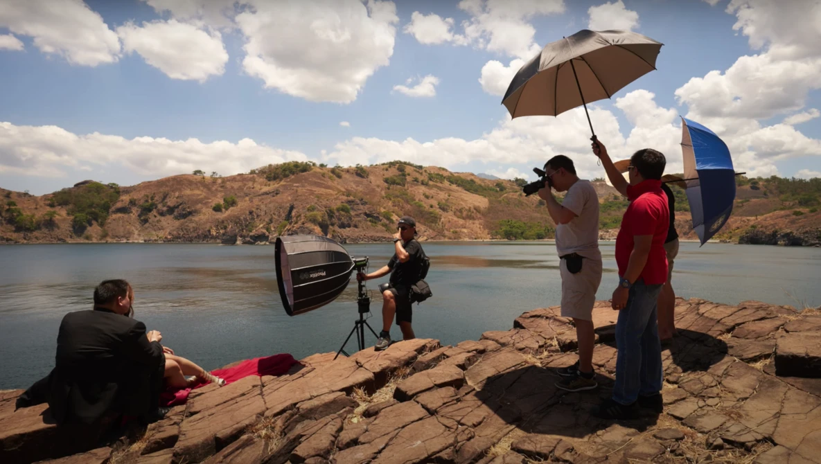 Film crew shooting by a rocky shoreline with umbrellas shading equipment and talent under bright daylight.