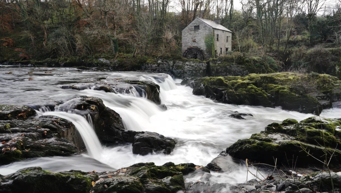 Rushing whitewater cascades over dark rocks in a river with a stone mill building nestled among bare trees on the bank.