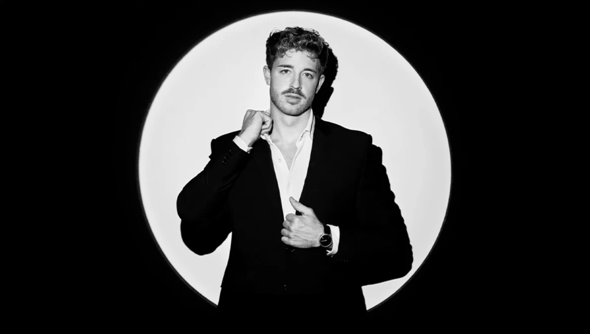 Man in black blazer and white shirt poses against circular white backdrop with confident expression.