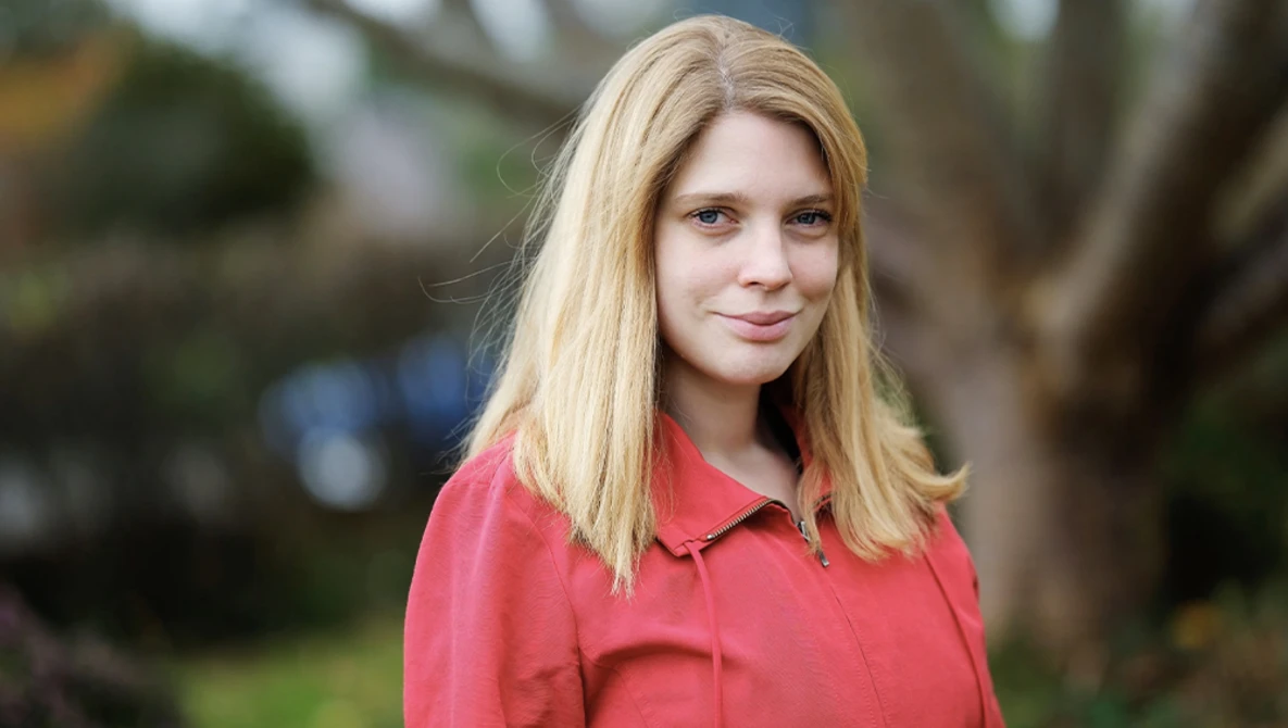 Blonde woman in coral pink shirt smiling outdoors with blurred greenery background.