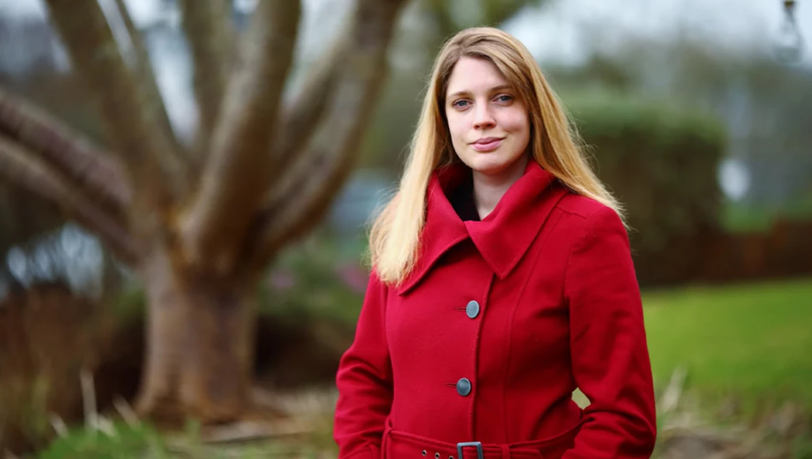 Young woman with long blonde hair wearing a bright red coat, photographed outdoors with soft natural lighting.