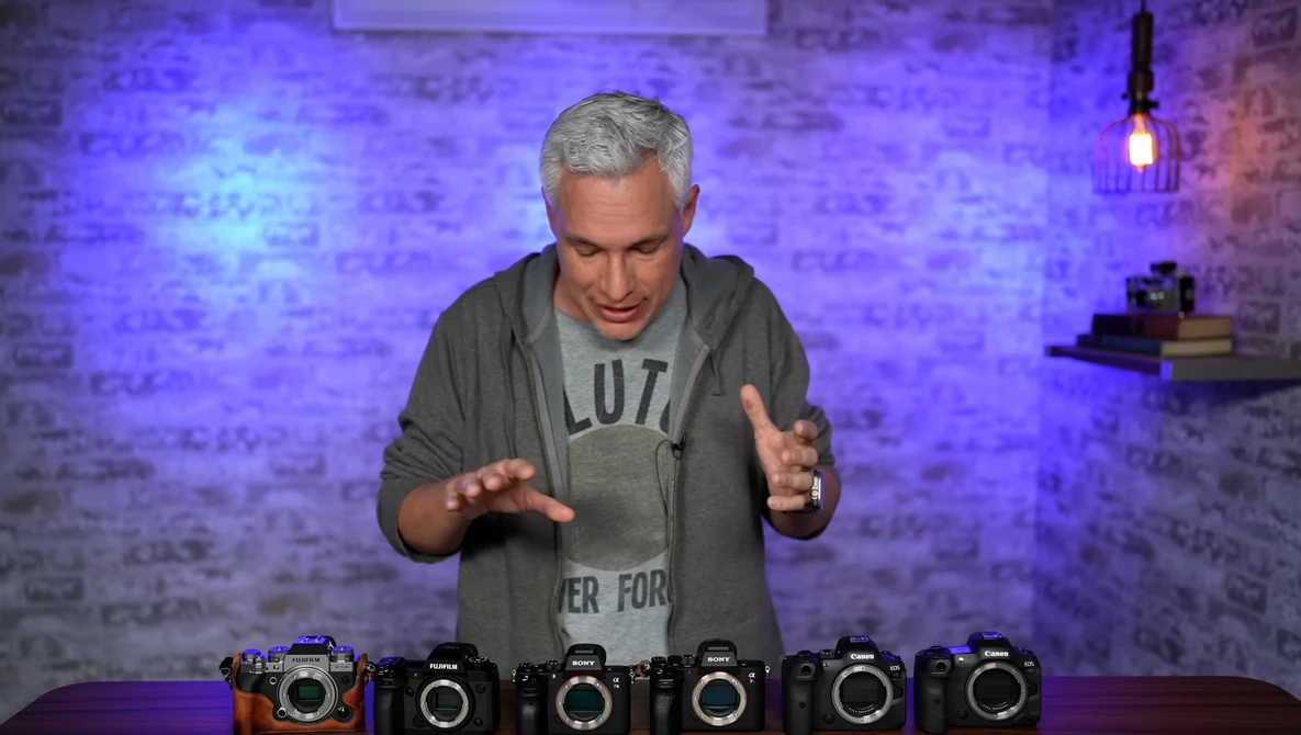 Man demonstrating six camera bodies arranged in a row on a table with purple brick wall backdrop.