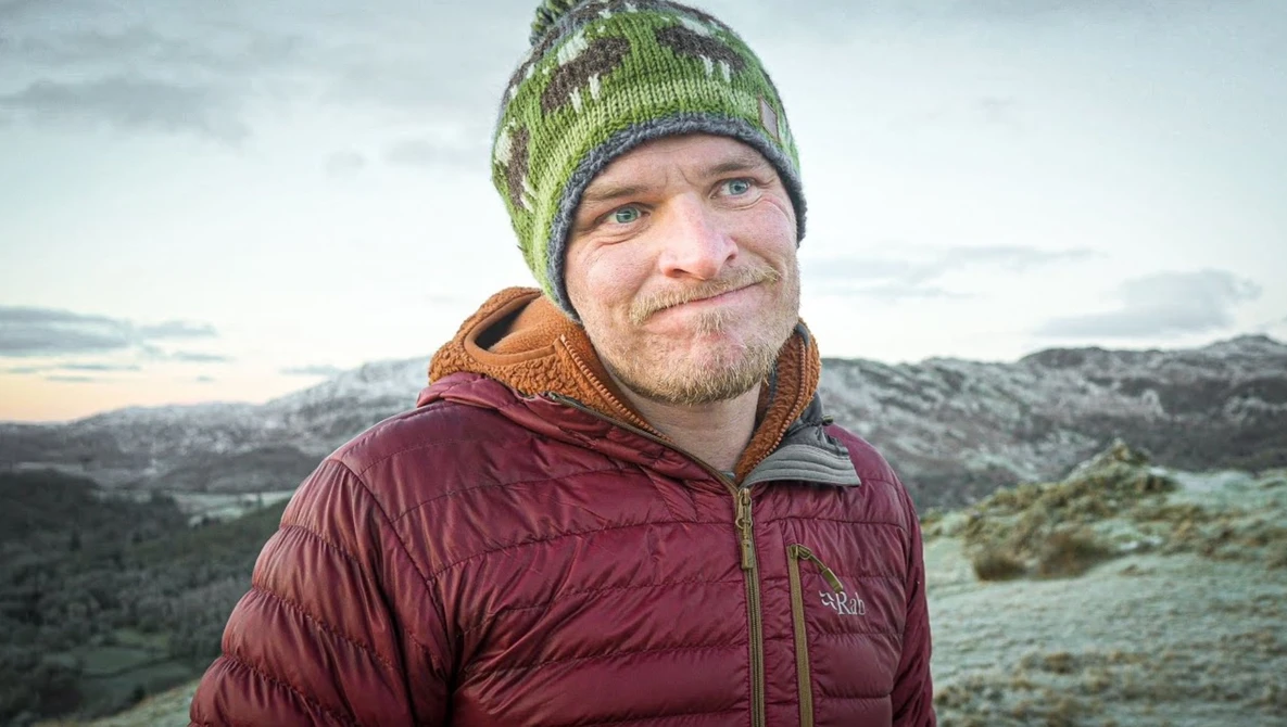 Man wearing maroon down jacket and green knit beanie in mountainous landscape at dusk.