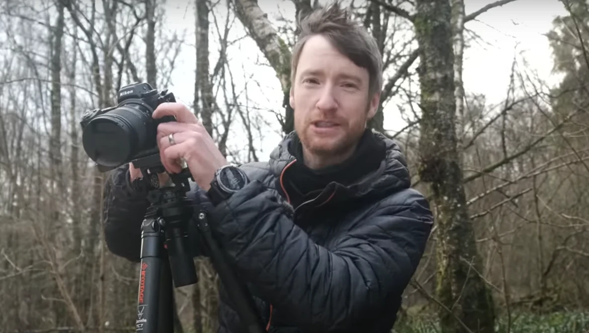 Male photographer holding a DSLR camera on a tripod in a bare winter forest.