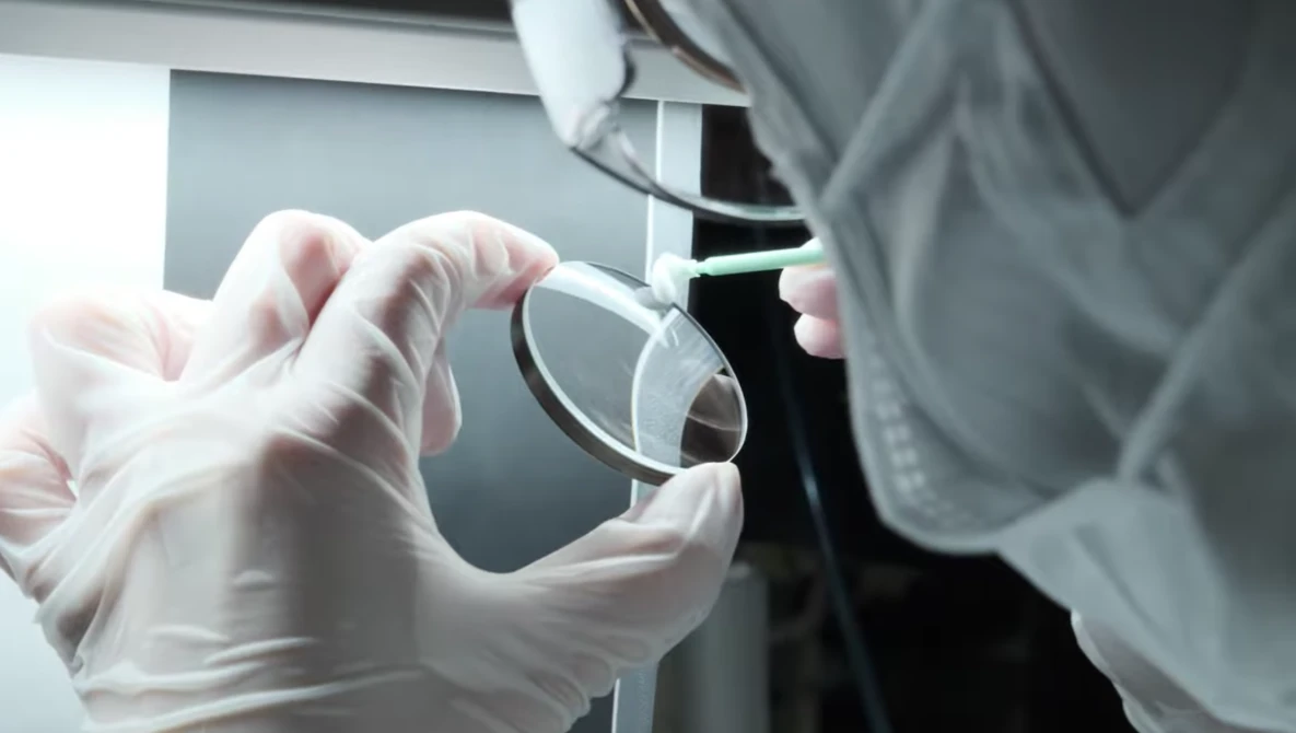 Gloved hands holding a clear glass lens or optical element during laboratory examination.
