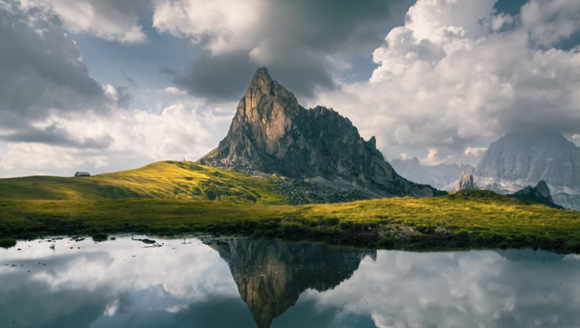 Dramatic alpine peak reflected in still water, surrounded by green meadow and dramatic cloudy sky.