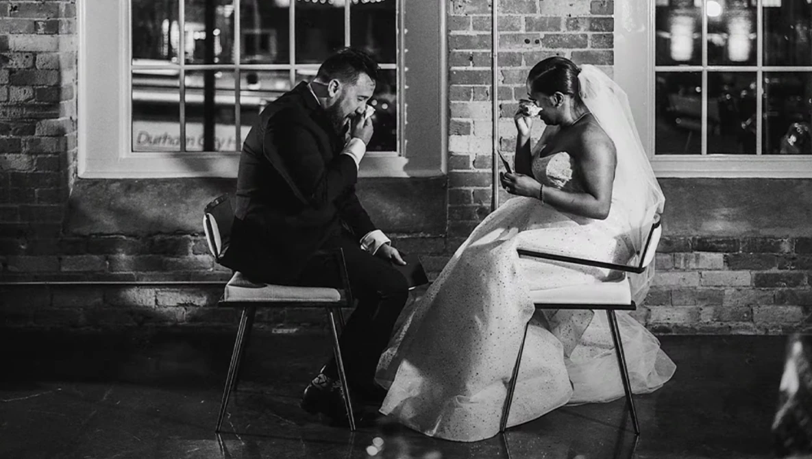Bride and groom sitting facing each other on chairs against a brick building with windows, captured in black and white photography.
