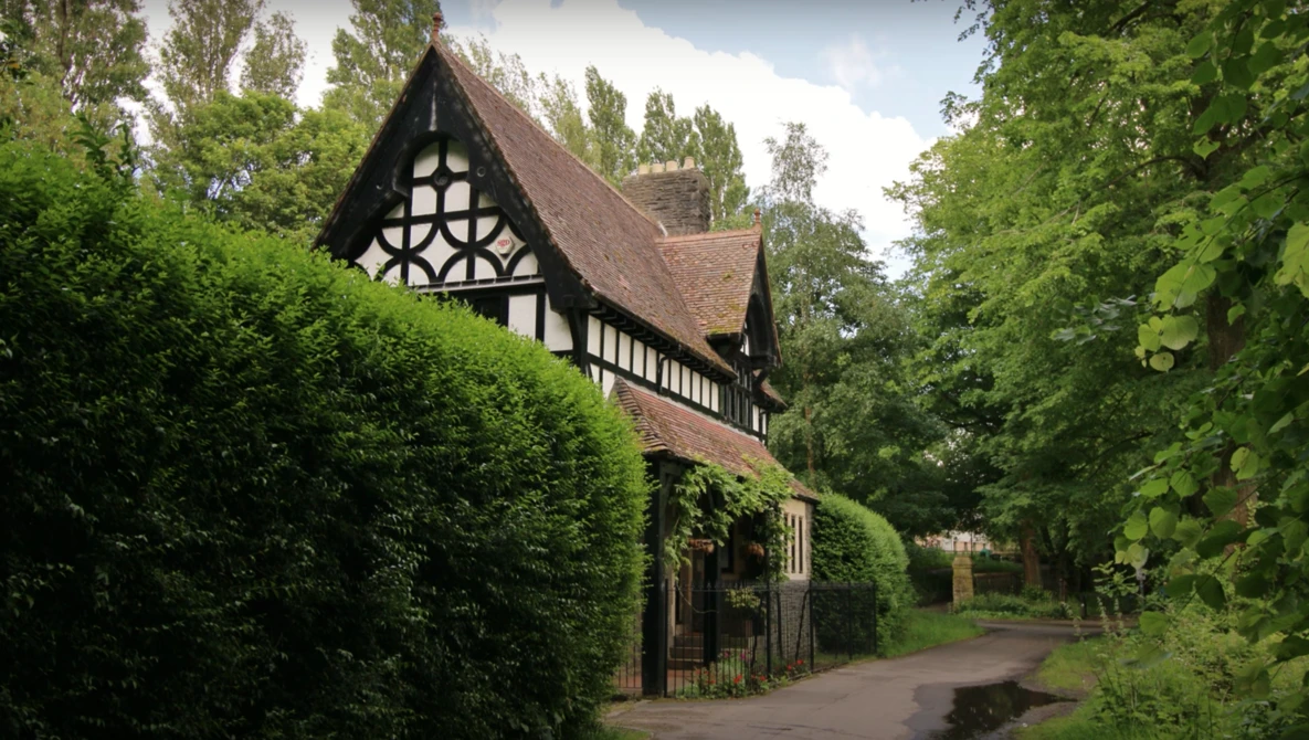 Half-timbered Tudor-style cottage surrounded by lush green hedges and trees.