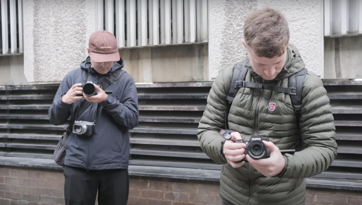 Two photographers holding compact cameras outside a modern building with corrugated metal siding.