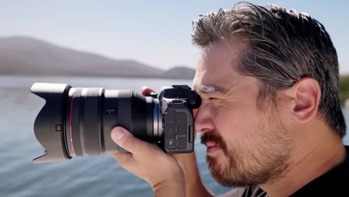 Photographer holding a mirrorless camera with telephoto lens to his eye, shooting toward a calm lake with mountains in the background.