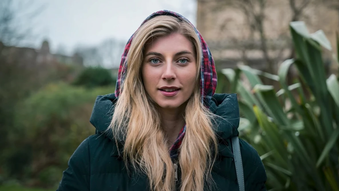 Woman with blonde hair wearing a dark jacket and plaid hood in an outdoor garden setting.