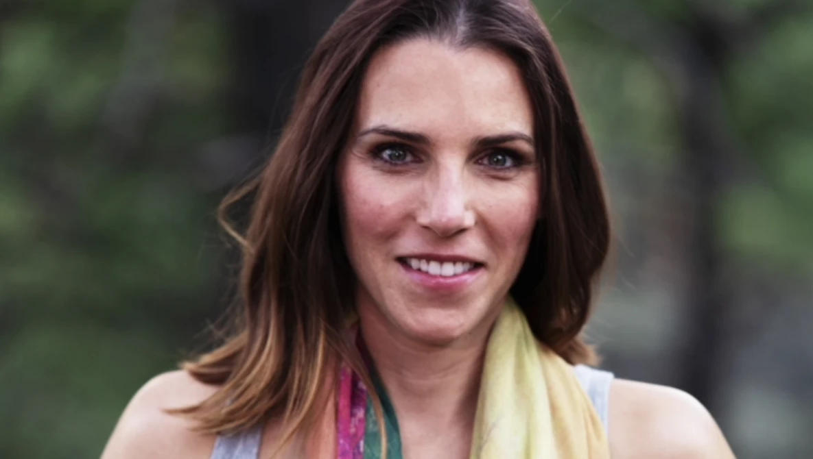 Woman with shoulder-length brown hair smiling at camera outdoors with blurred green foliage background.
