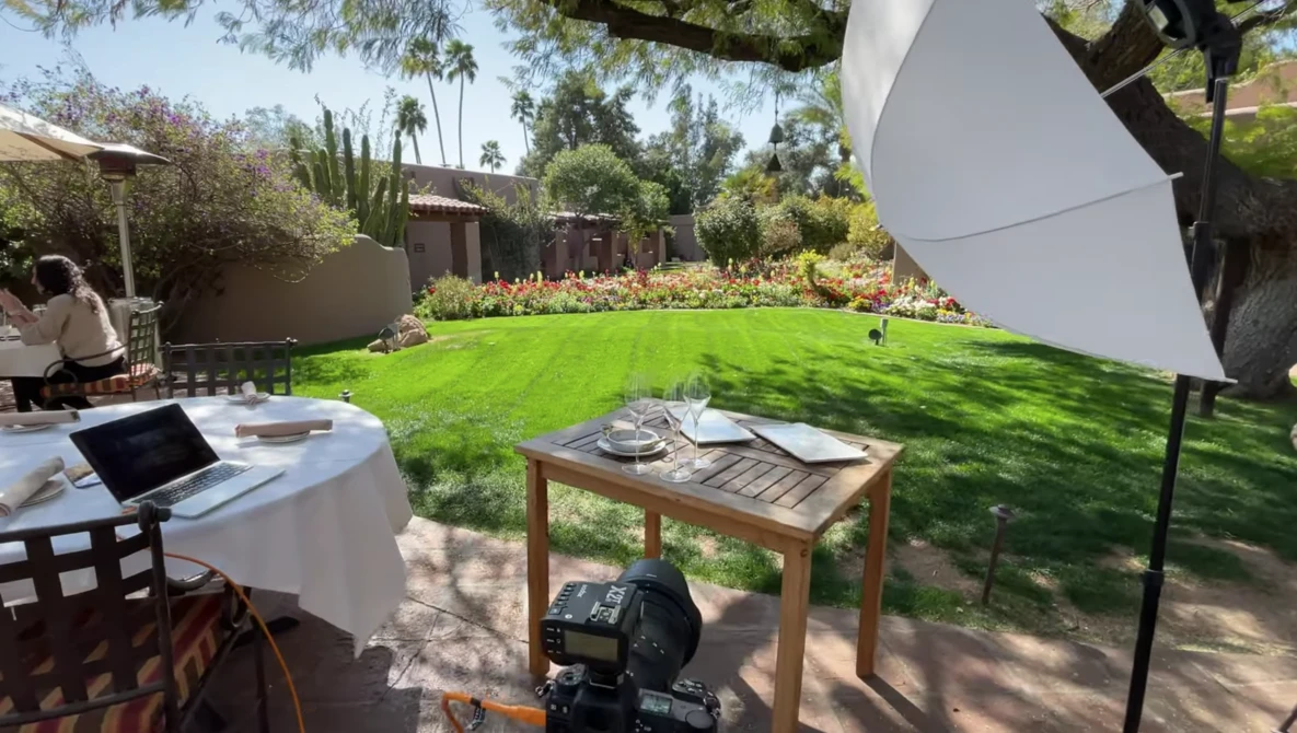 Outdoor photography setup in a sunlit garden with large reflector, wooden table, and shade sail.