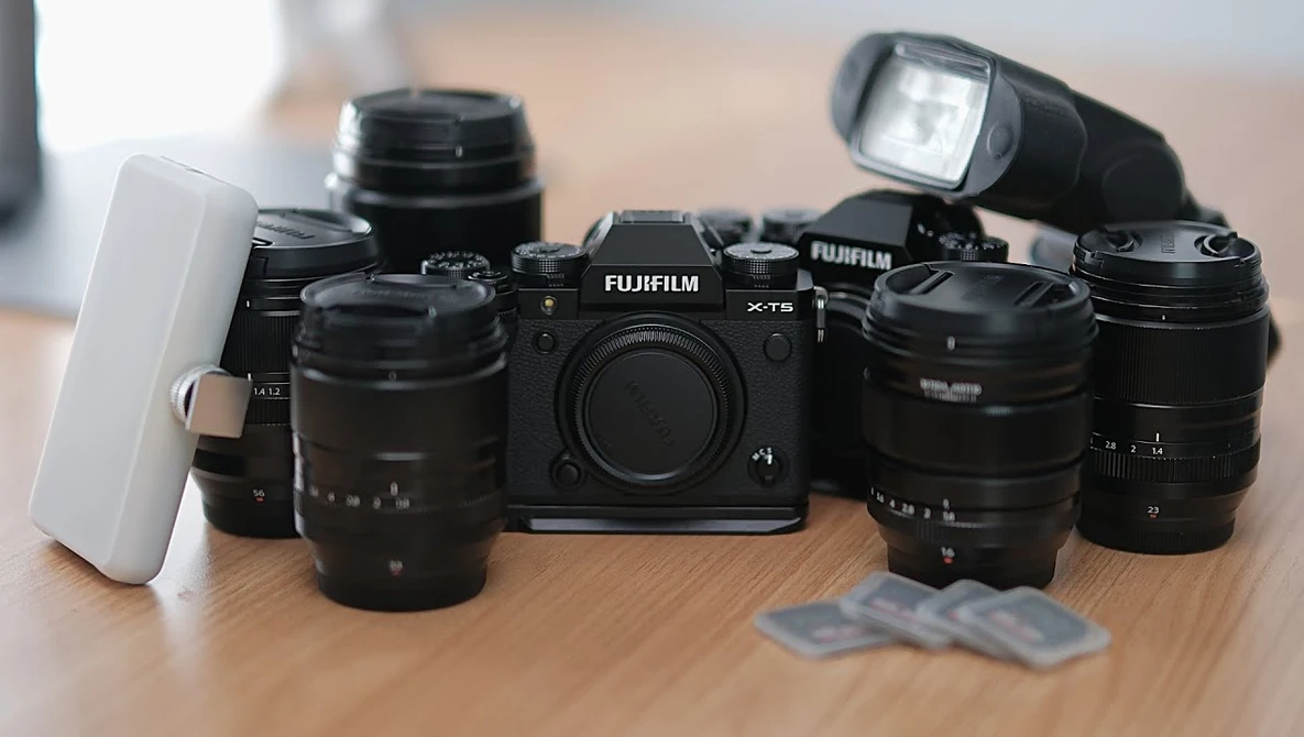Fujifilm X-T5 camera body surrounded by multiple black lenses, a white diffuser, and external flash on wooden surface.