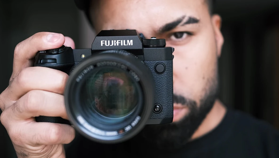 Photographer holding a Fujifilm mirrorless camera with telephoto lens pointed toward camera.