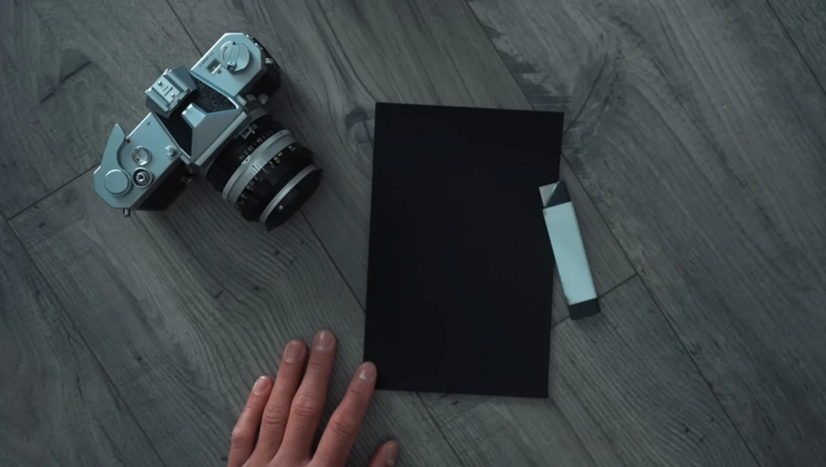 Flat lay of vintage rangefinder camera with lens, black notebook, and photographer's hand on wooden surface.