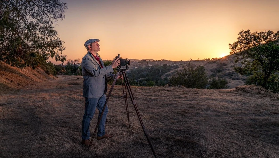 Surveyor in business casual attire operating a tripod-mounted surveying instrument during golden hour on a hillside.