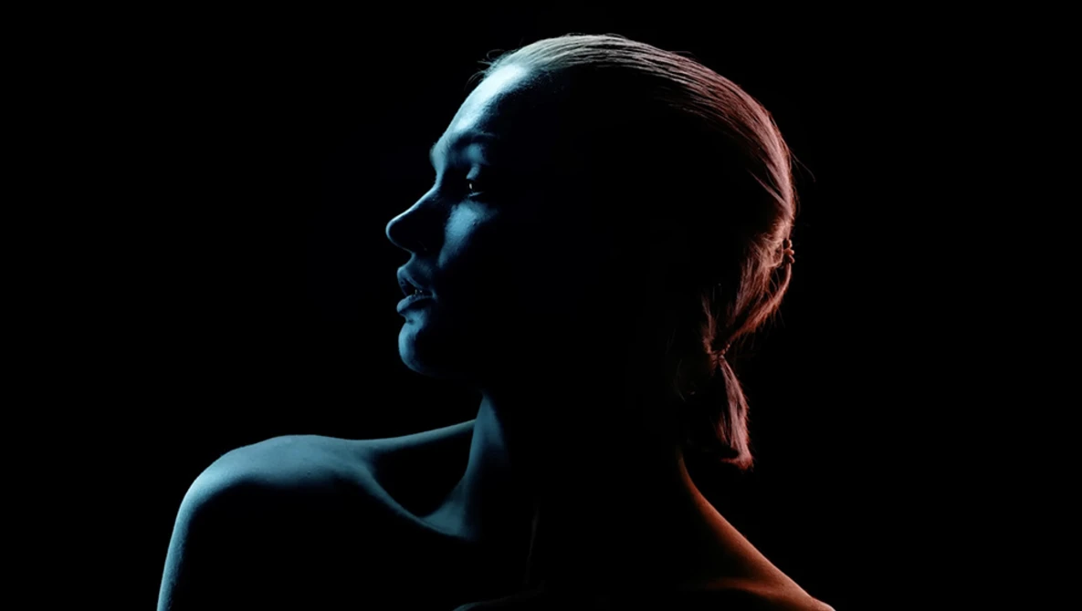 Profile of a woman's face and shoulders against a black background with warm and cool colored rim lighting.